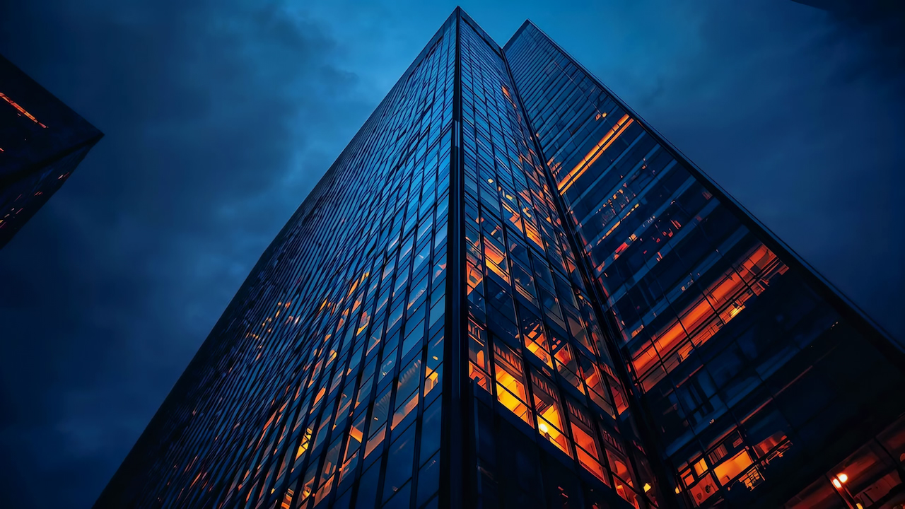 A blue glass tower at dusk, its lit floors seen from below